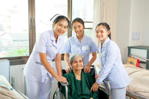 ผู้สูงอายุหญิงนั่งบนวีลแชร์ โดยมีพยาบาลหญิง และทีมบริบาลยืนล้อมรอบ ทุกคนยิ้มอย่างมีความสุข
