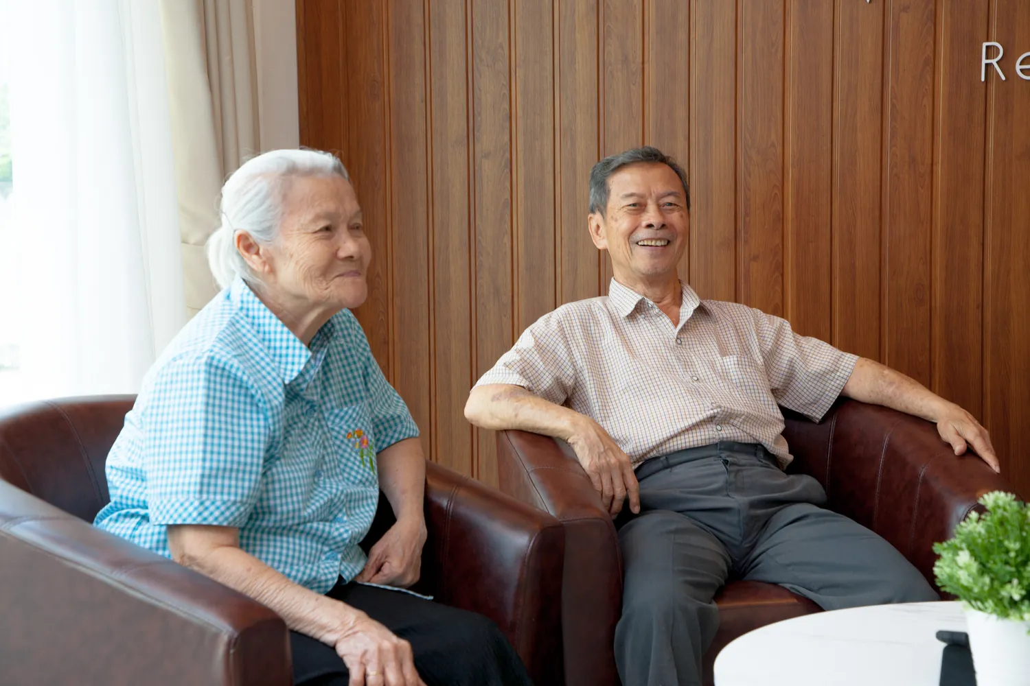ผู้สูงอายุหญิงนั่งอยู่ข้างผู้สูงอายุชาย โดยนั่งยิ้มอยู่ที่โซฟาภายในศูนย์ดูแลผู้สูงอายุวินเนสต์
