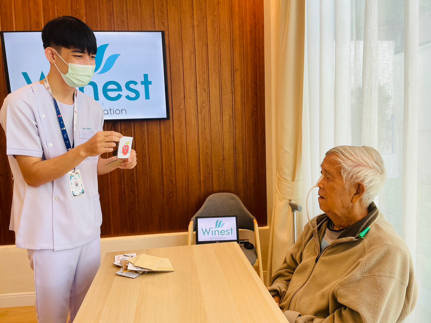 บุรุษพยาบาลจัดกิจกรรมโชว์ภาพผลไม้ ให้กับผู้สูงอายุชาย ที่ใส่สายให้อาหารทางจมูก