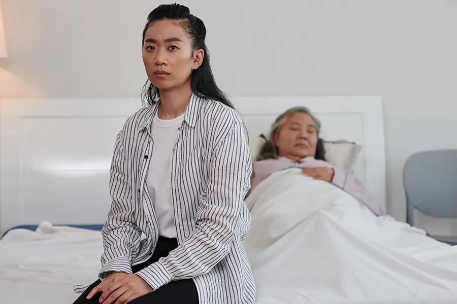ผูู้ดูแลหญิงชาวเอเชียสีหน้าเหนื่อยล้า นั่งอยู่ปลายเตียงที่มีผู้สูงอายุหญิงนอนหลับอยู่